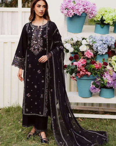 Woman in a black embroidered outfit standing in front of a white fence with potted flowers.