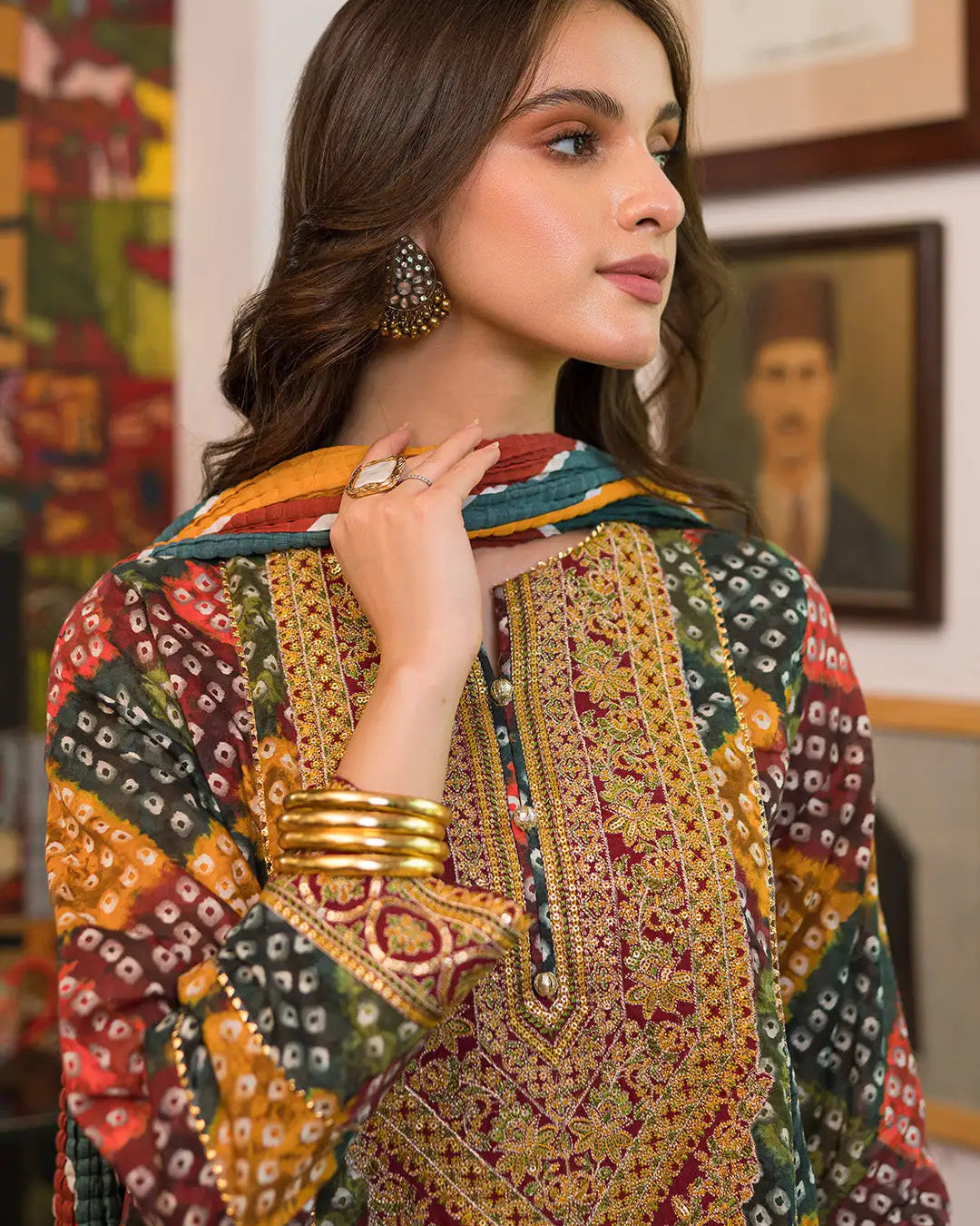 A woman wearing a colorful embroidered traditional outfit with a patterned dupatta, gold bangles, and statement earrings, posed indoors against a wall with framed art.