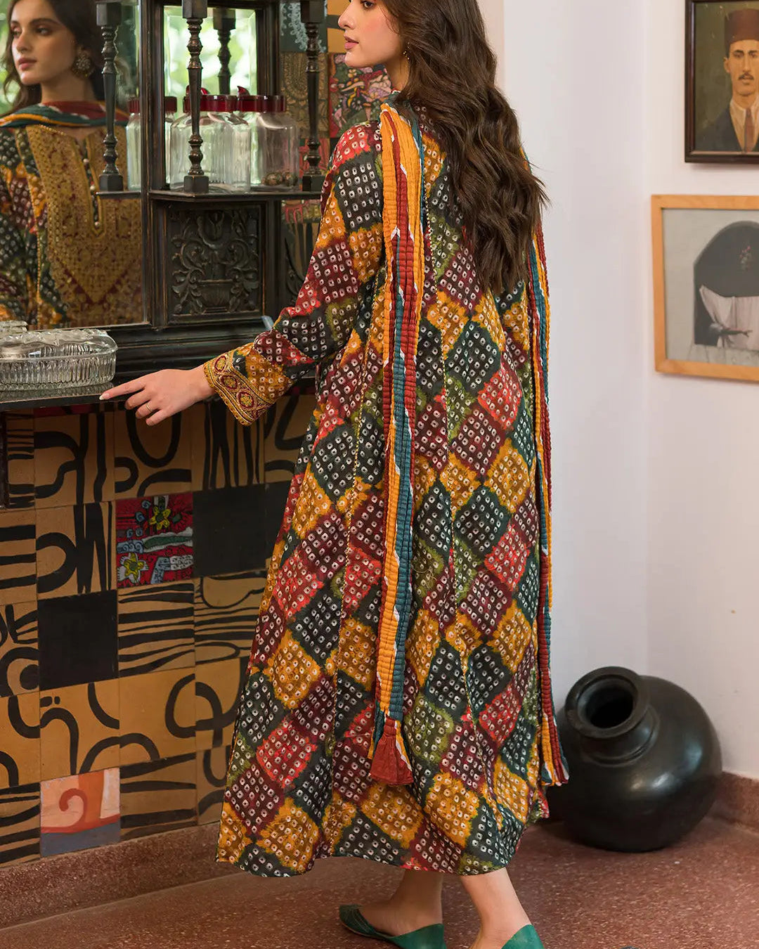 A woman wearing a long-sleeved, knee-length kurta with a colorful geometric block print pattern in yellow, red, green, and black, standing in an indoor setting with a mirror and decorative wall art.