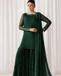 A woman models a dark green traditional ethnic outfit consisting of a long-sleeve kurta, matching flared pants, and a sheer dupatta with embroidered borders, set against a neutral draped curtain background.