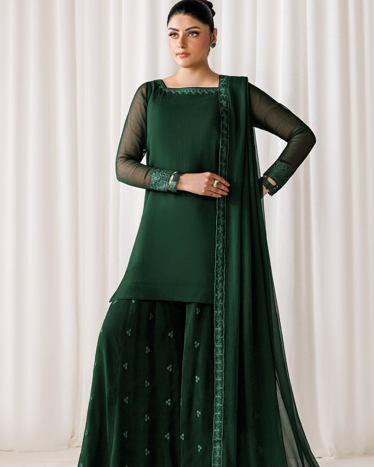 A woman models a dark green traditional ethnic outfit consisting of a long-sleeve kurta, matching flared pants, and a sheer dupatta with embroidered borders, set against a neutral draped curtain background.