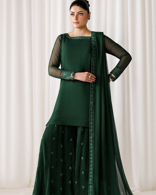 A woman models a dark green traditional ethnic outfit consisting of a long-sleeve kurta, matching flared pants, and a sheer dupatta with embroidered borders, set against a neutral draped curtain background.