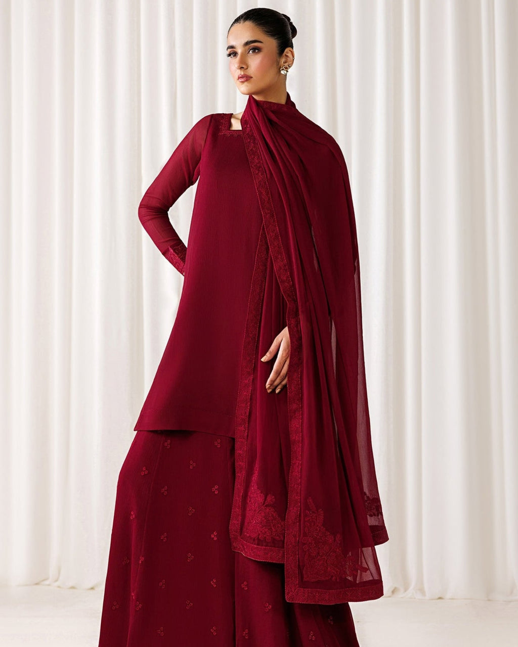 A woman models a deep maroon red traditional outfit consisting of a long-sleeve kurta, matching wide-leg pants, and a sheer draped scarf, set against a soft white curtain background.