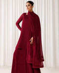 A woman models a deep maroon red traditional outfit consisting of a long-sleeve kurta, matching wide-leg pants, and a sheer draped scarf, set against a soft white curtain background.