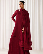 A woman models a deep maroon red traditional outfit consisting of a long-sleeve kurta, matching wide-leg pants, and a sheer draped scarf, set against a soft white curtain background.