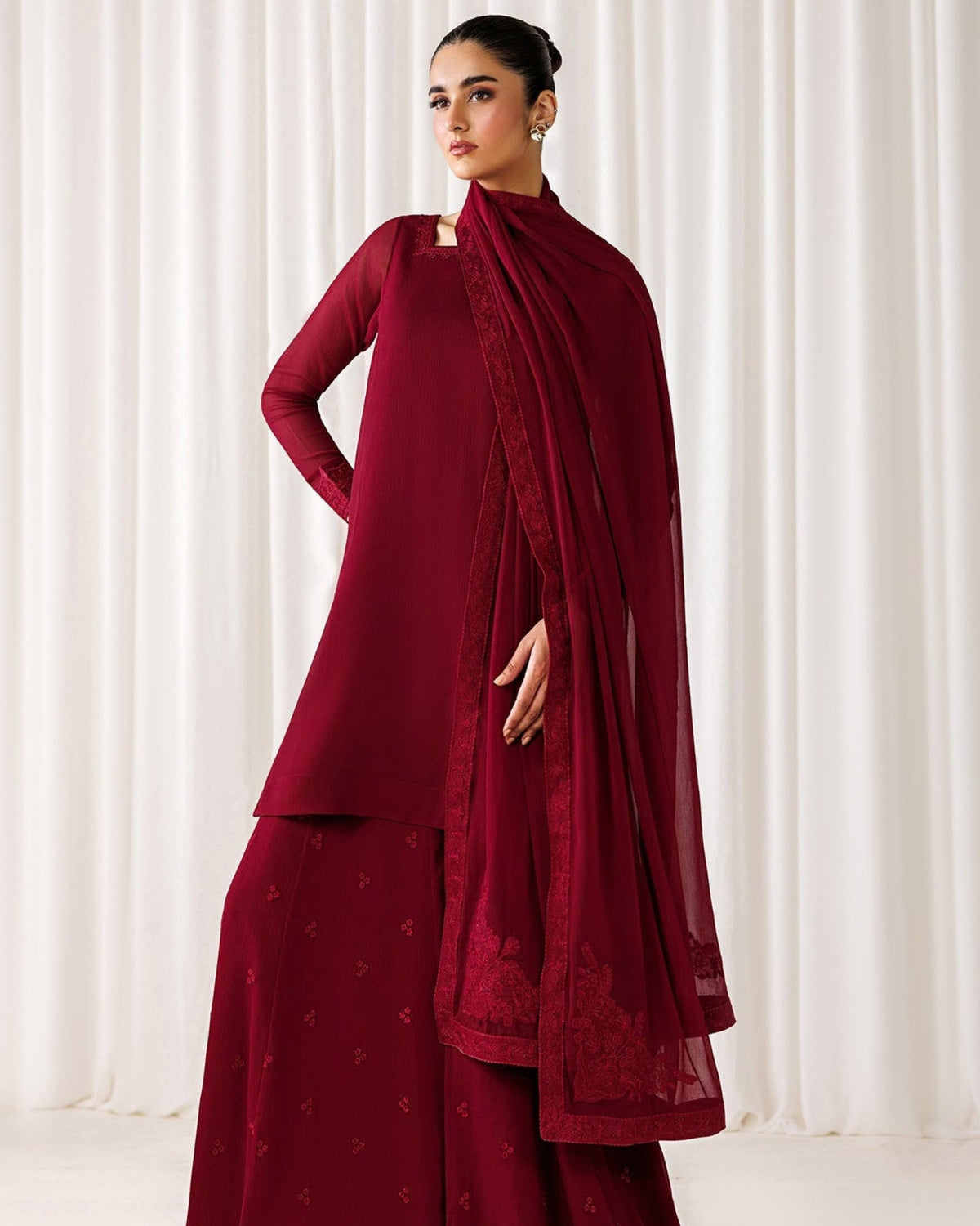 A woman models a deep maroon red traditional outfit consisting of a long-sleeve kurta, matching wide-leg pants, and a sheer draped scarf, set against a soft white curtain background.