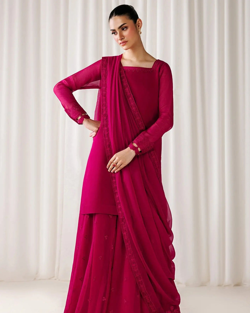 A woman models a magenta pink traditional Indian outfit consisting of a long-sleeve kurta, matching flared pants, and a draped dupatta, set against a soft white curtain background.