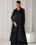 A woman models a black traditional outfit consisting of a kurta, wide-leg pants, and a matching sheer embroidered cardigan, set against a neutral cream-colored curtain backdrop.