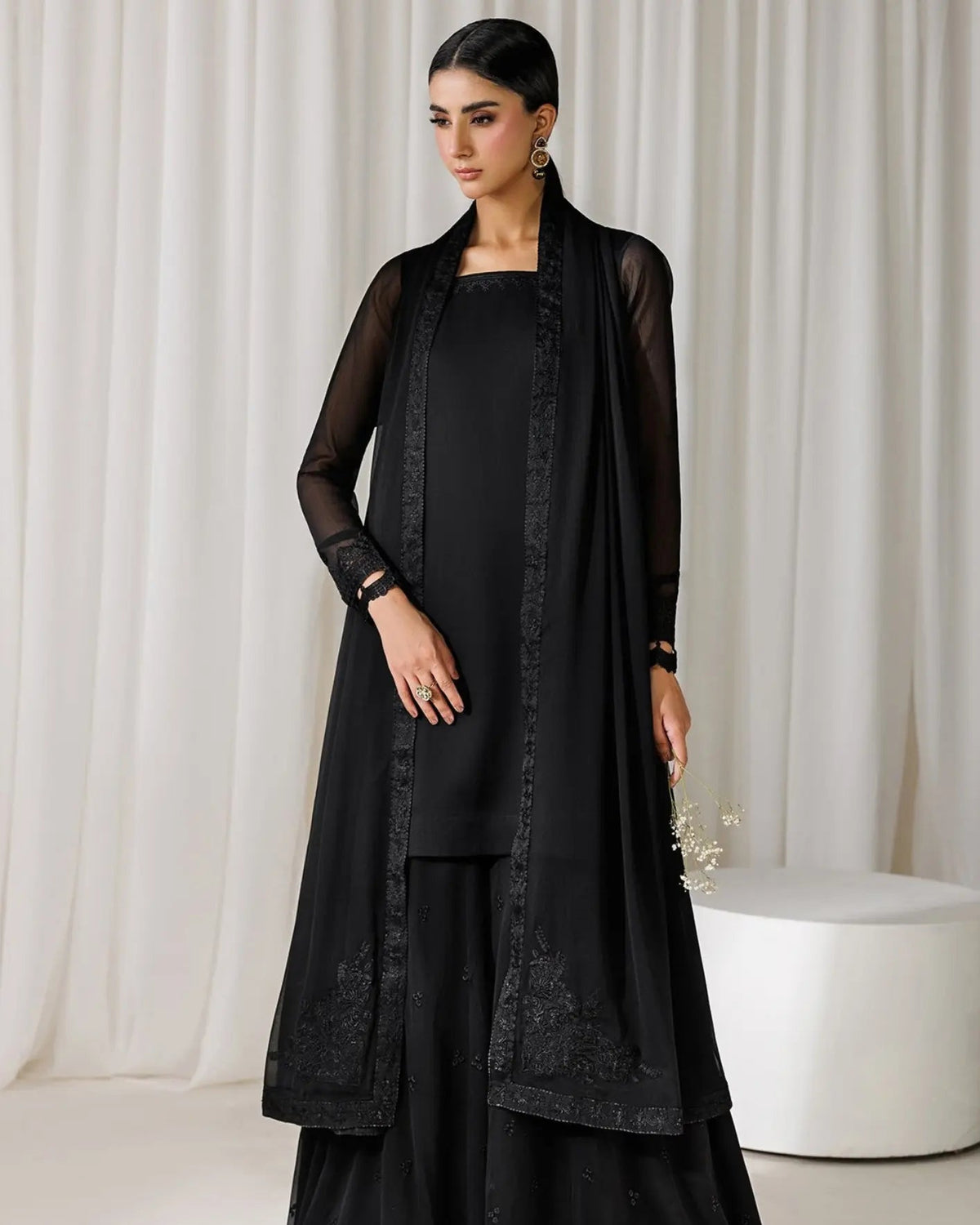 A woman models a black traditional outfit consisting of a kurta, wide-leg pants, and a matching sheer embroidered cardigan, set against a neutral cream-colored curtain backdrop.