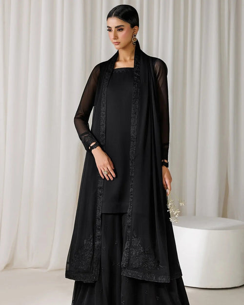 A woman models a black traditional outfit consisting of a kurta, wide-leg pants, and a matching sheer embroidered cardigan, set against a neutral cream-colored curtain backdrop.