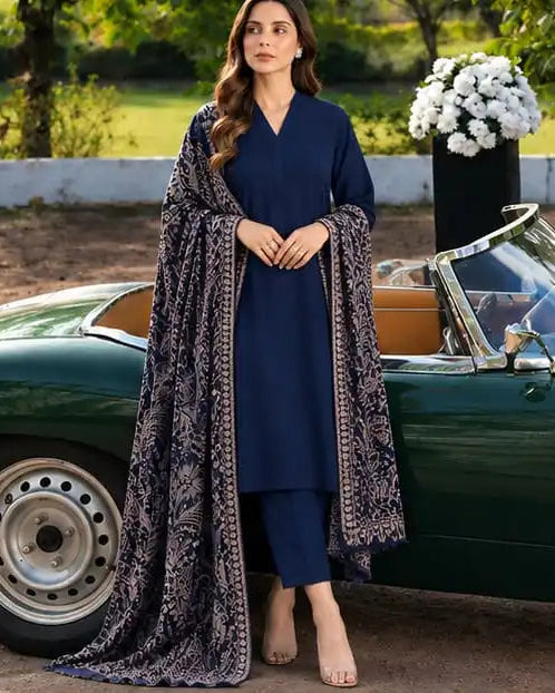 A woman stands outdoors beside a green vintage convertible car, wearing a navy blue kurta set with a long patterned shawl draped over her shoulders. The setting includes trees, a fence, and a flower arrangement in the background.