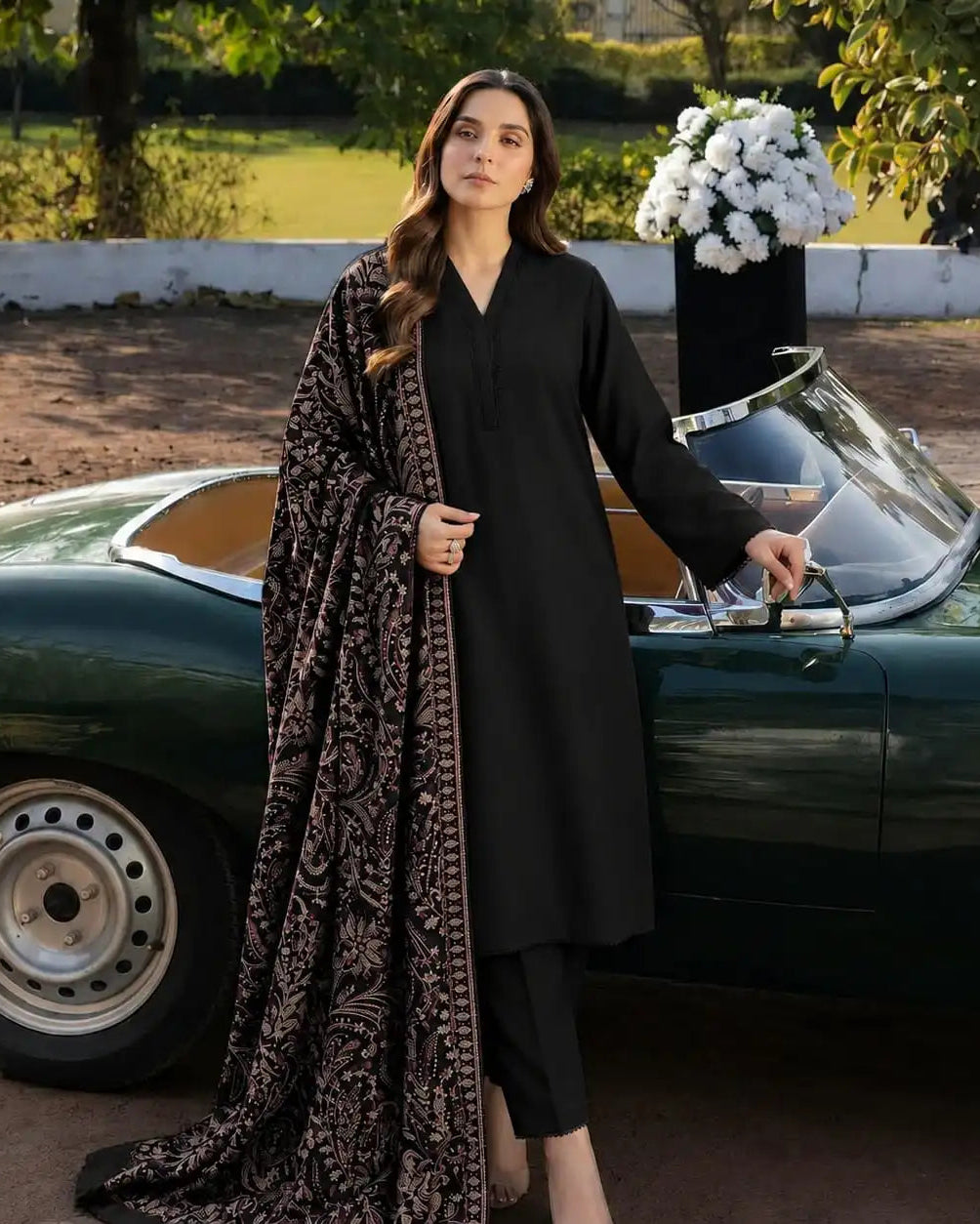 A woman stands outdoors beside a dark green vintage convertible car, wearing a black kurta with black pants and a long patterned dupatta draped over one shoulder. The background includes trees, a white barrier, and a vase of white flowers.