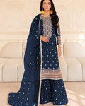 A woman models a navy blue traditional outfit with intricate silver and gold embroidery, featuring a long kurta, flared pants, and a matching dupatta, posed in front of a cream-colored sofa.
